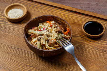 Wok udon noodles with beef and vegetables in a coconut shell plate on a napkin with a fork next to sesame seeds and soy sauce. Horizontal photo.の写真素材
