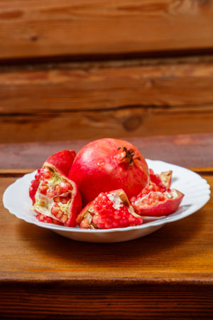 Fresh pomegranate broken with grains and a whole fruit on a white plate on a wooden table. Vertical photoの写真素材