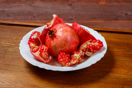 Fresh pomegranate broken with grains and a whole fruit on a white plate on a wooden table Horizontal photoの写真素材