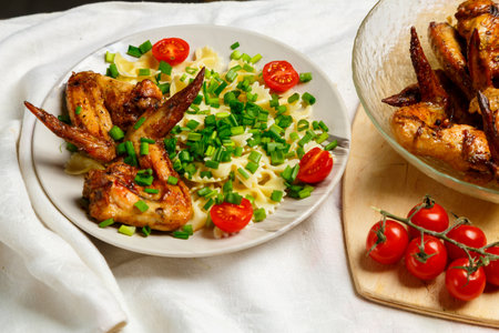 Farfalle pasta with grilled chicken wings on a white plate decorated with cherry tomatoes and green onions next to a dish with wings. Medium plan Horizontal photoの写真素材
