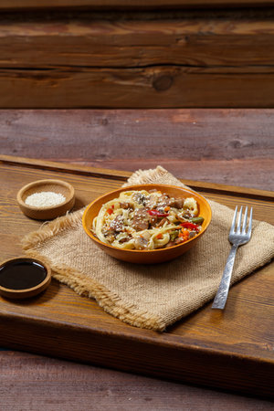 Wok udon noodles with beef and vegetables in a wooden bowl on a salad on a hearth board next to soy sauce and sesame seeds. Vertical photo.の写真素材