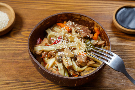 Wok udon noodles with beef and vegetables in a coconut shell plate on a napkin with a fork next to sesame seeds and soy sauce. Horizontal photo.の写真素材