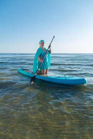 A woman in a turquoise swimsuit with a skirt and a scarf on her head on a SUP board near the sea. Vertical photoの写真素材
