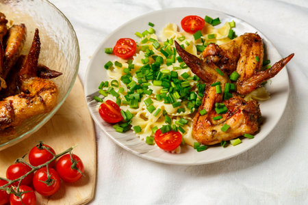 Farfalle pasta with grilled chicken wings on a white plate decorated with cherry tomatoes and green onions next to a dish with wings. horizontal photoの写真素材