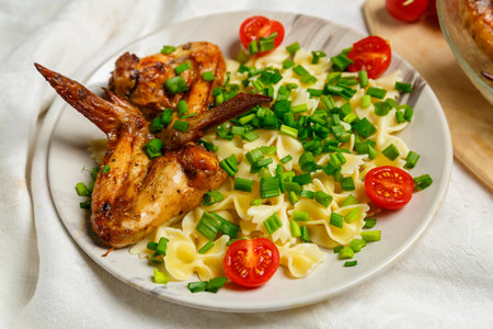 Farfalle pasta with grilled chicken wings on a white plate on a salad garnished with cherry tomatoes and green onions. Medium planの写真素材
