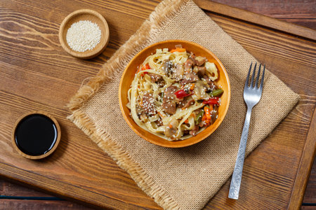 Wok udon noodles with beef and vegetables in a wooden plate on a salad on a hearth board next to soy sauce and sesame seeds. Horizontal photo.の写真素材
