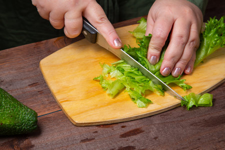 A woman's hand cuts green leaves with a knife on a wooden board. Horizontal photo.の写真素材