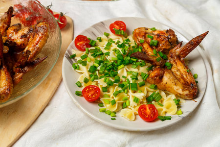 Farfalle pasta with grilled chicken wings on a white plate decorated with cherry tomatoes and green onions next to a dish with wings. Medium plan.の写真素材