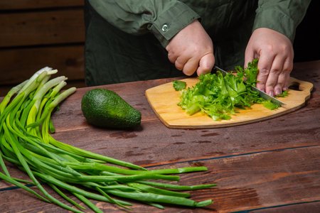 A woman's hand cuts greens in half with a knife on a wooden board on a table with vegetables. Horizontal photo.の写真素材