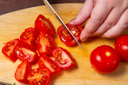 A woman's hand cuts a cherry tomato with a knife on a wooden board on the table. horizontal photoの写真素材