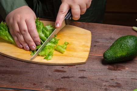 A woman's hand cuts greens in half with a knife on a wooden board on a table with vegetables. Horizontal photo.の写真素材