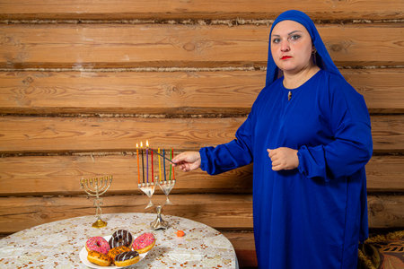 A Jewish woman with a blue cap and veil on her head lights Hanukkah candles at a table with donuts, Horizontal photo.の写真素材