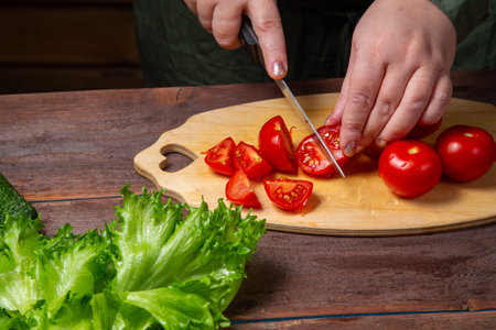 A woman's hand cuts a cherry tomato with a knife on a board on a table with vegetables. horizontal photoの写真素材