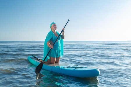 A Jewish woman in a turquoise swimsuit with a skirt and a scarf on her head kneeling on a SUP board near the sea. horizontal photoの写真素材