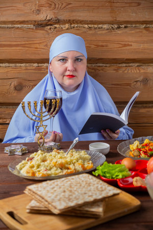 A Jewish woman in a blue head veil reads kiddush at the Pesach Seder table with a glass of wine....