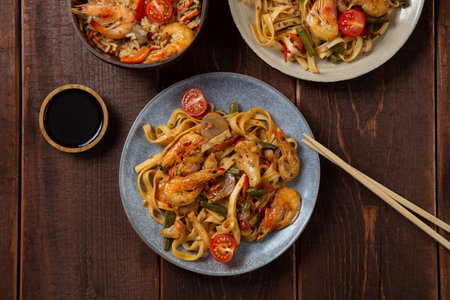 Rice with shrimp and udon in yaki soba sauce on a wooden table next to cherry tomatoes, soy sauce and chopsticks. horizontal photoの写真素材