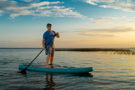 A man in shorts and a T-shirt swims on a lake on a paddle board against the backdrop of a sunset sky with clouds. Horizontal photo.の写真素材