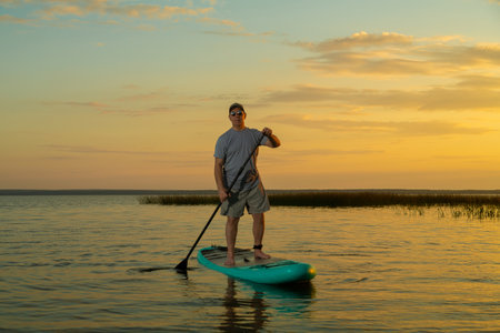 A man in shorts and a T-shirt swims on a lake on a stand-up paddle board at sunset. Horizontal...