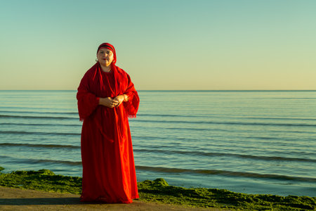 Muslim woman in red dress and headscarf standing on seashore at dawn in the morning. Horizontal...