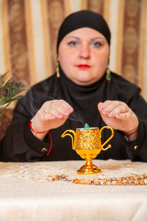 A Muslim woman in a black hijab makes a dua at a table with incense. Vertical photo.の写真素材