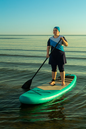 A woman in a modest Islamic burkini on a paddleboard with a raised paddle in the sea against the backdrop of the dawn sky. Vertical photo.の写真素材