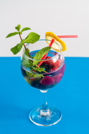 Non-alcoholic cocktail raspberries blueberries lemon and mint in a glass with a straw on a blue background. Vertical photoの写真素材