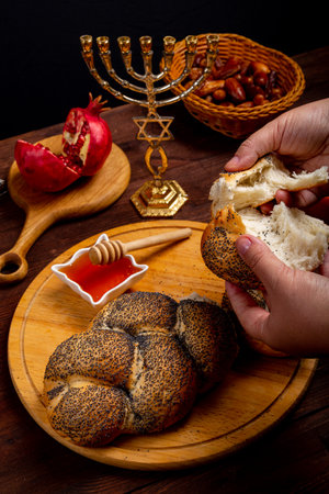 Female hands breaking challah on the table meal in Rosh Hashanah next to apple honey menorah burning candles. Vertical photo.の写真素材