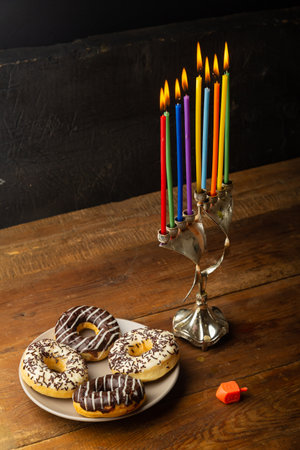 Hanukkiah with colored candles on the table and chocolate glazed donuts and dreidel. Vertical photo.の写真素材