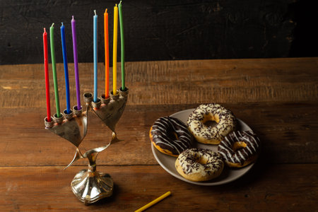 Hanukkiah with candles on the table in the semi-darkness next to glazed donuts and dreidel. Horizontal photo.の写真素材