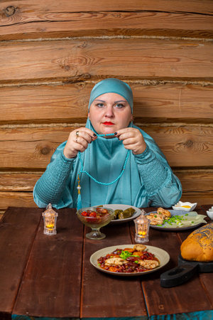 Muslim woman in blue hijab at set table during Ramadan reading dhikr fingering beads. Vertical photoの写真素材