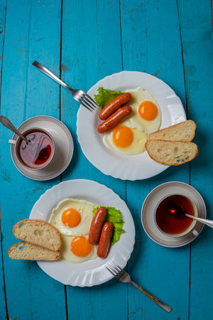 Breakfast for two fried eggs with sausages, fresh croutons, and tea in a cup on a blue wooden table. Vertical photo.の写真素材