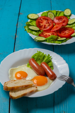 Salad, cucumbers and tomatoes in olive oil and fried eggs with sausages in a plate on a blue wooden table. Vertical photoの写真素材