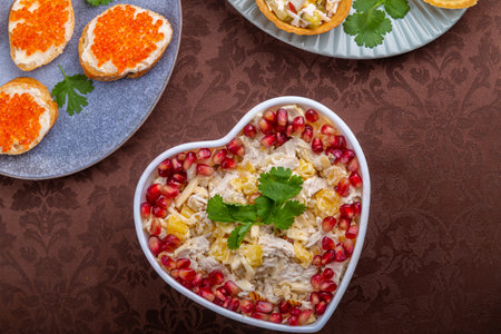 Salad with chicken and pomegranates in a heart-shaped salad bowl next to tartlets and sandwiches with caviar on a brown tablecloth. horizontal photoの写真素材