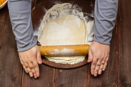 Women's hands roll out dough with a rolling pin on a wooden table on a round baking board for Purim....