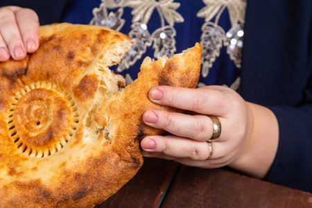 A Muslim woman without a face in a blue hijab and embroidered national dress at a set table with food Eid al fitr breaks a flatbread close-up. Horizontal photo.の写真素材