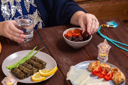 A Muslim woman without a face in a blue hijab and embroidered national dress at a set table with food Eid al fitr eats a date and water. Horizontal photo.の写真素材