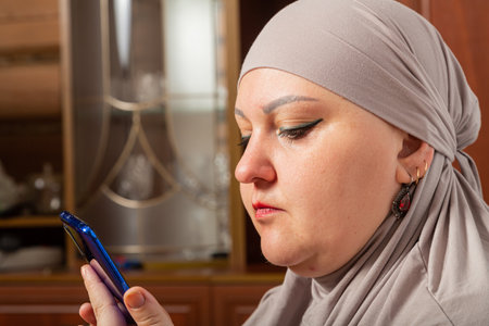 Muslim woman in gray hijab in profile in room chatting on smartphone social network close-up. Horizontal photo.の写真素材