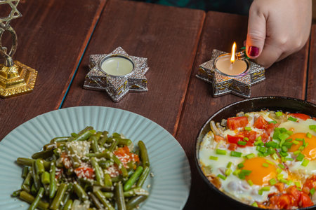 A Jewish woman's hand lights Shabbat candles at a table with festive dishes. Horizontal photo.の写真素材