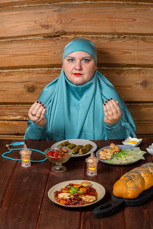 Muslim woman in blue hijab with table set in Ramadan making dua before iftar. Vertical photoの写真素材