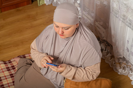 Muslim woman in gray hijab in room on plu on pillows scrolls corresponds on smartphone. Horizontal photo.の写真素材
