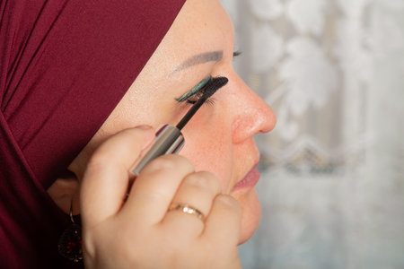 Muslim woman in red hijab sits in room at mirror and applies make-up mascara close-up. Horizontal...