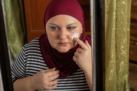 Muslim woman in red hijab sits in room near mirror and applies cream on face close-up. Horizontal photo.の写真素材
