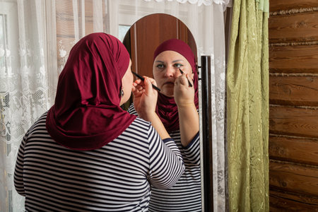 Muslim woman in red hijab near mirror and applying cream on face medium shot. Horizontal photo.の写真素材