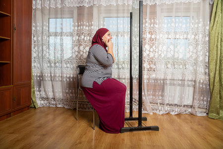 Muslim woman in hijab sitting in the room at the mirror and applying makeup, general plan. Horizontal photo.の写真素材