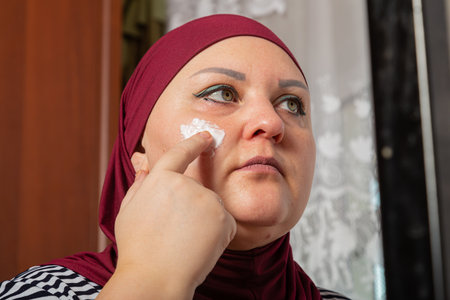 Muslim woman in red hijab near mirror and applying cream on face close-up. Horizontal photo.の写真素材