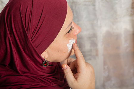 Muslim woman in red hijab sits in room near mirror and applies cream on face close-up. Horizontal photo.の写真素材