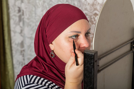 Muslim woman in red hijab sits in room near mirror and applies make-up close-up. Horizontal photo.の写真素材