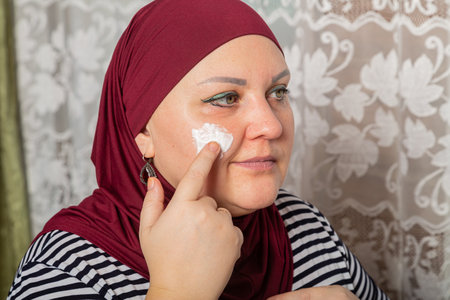 Muslim woman in red hijab sits in room near mirror and applies cream on face close-up. Horizontal photo.の写真素材