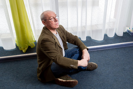 A middle-aged man in a green jacket sits on the floor near a panoramic window. Horizontal photo.の写真素材