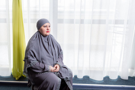 A Muslim woman in a modest gray hijab sits on a stool against the backdrop of a panoramic window. Horizontal photo.の写真素材